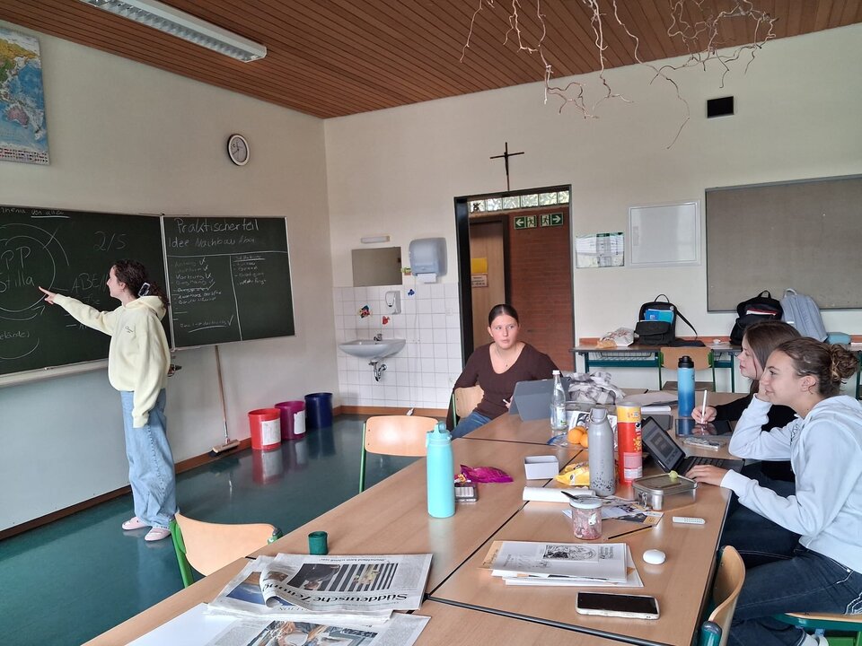 Schülerinnen arbeiten im Klassenzimmer an ihren Projektpräsentationen, eine erklärt etwas an der Tafel